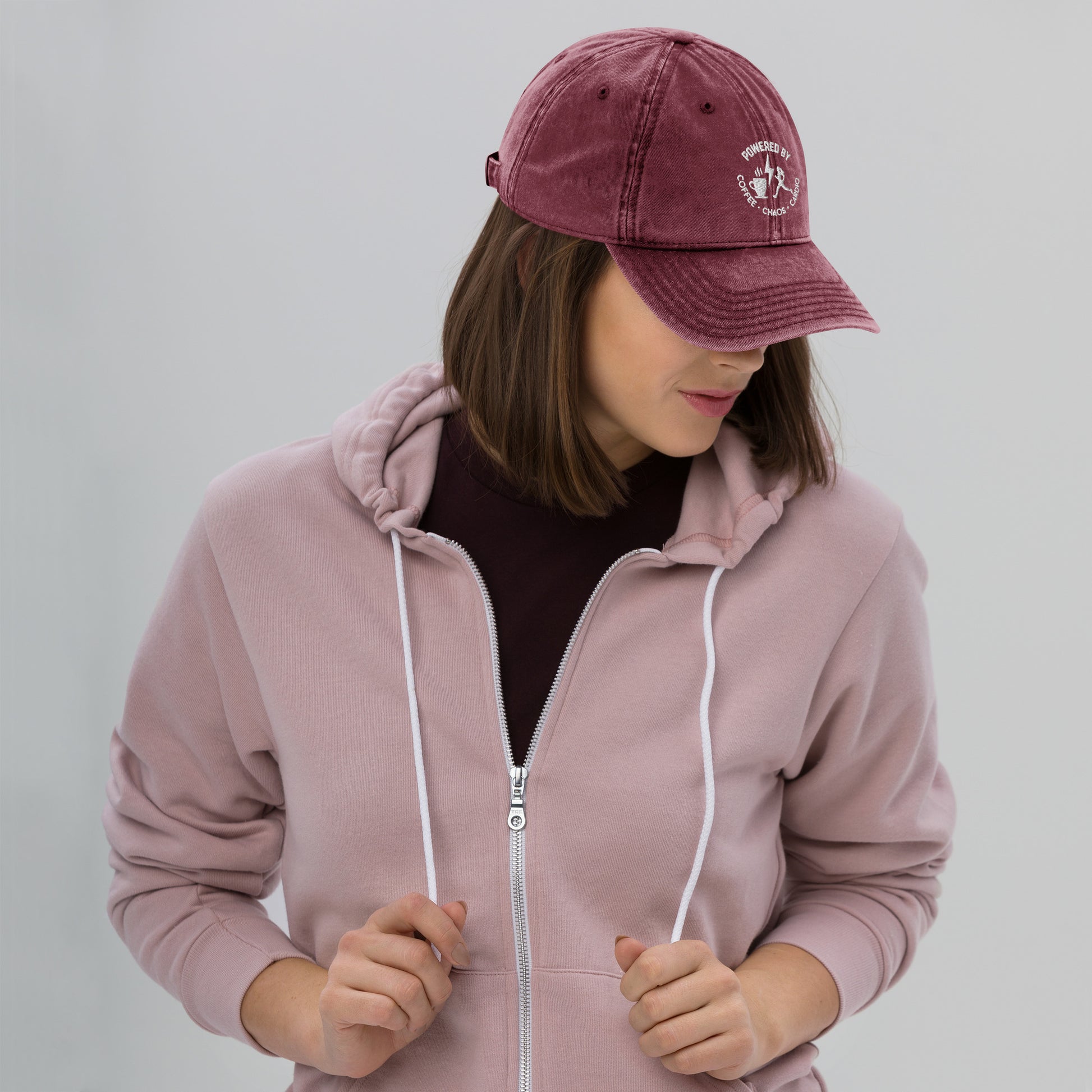 Maroon vintage cotton twill cap with white embroidered "Powered by Coffee, Chaos & Cardio" text worn by woman in mauve hoodie.